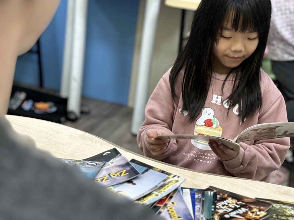 A young girl in a pink sweatshirt reading a picture book at a table.
